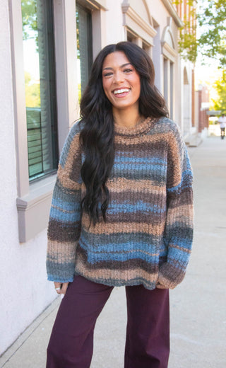 A woman smiling outdoors wearing a chunky knit sweater with horizontal stripes in blue, brown, beige, and gray, paired with dark purple pants.