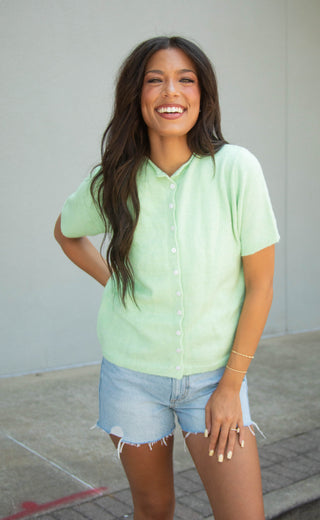 A woman smiling and wearing a light mint green short-sleeve button-up cardigan and light-wash denim shorts, standing outdoors against a plain light gray wall.