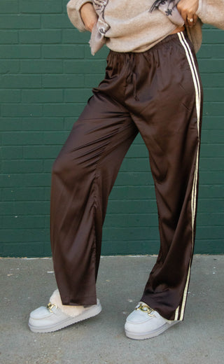 A person wearing dark brown satin-like wide-leg pants with a white side stripe, paired with white slip-on shoes, standing against a dark green brick wall.