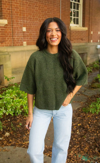A woman smiling outdoors in front of a brick building, wearing a loose olive green knit sweater and light blue jeans.
