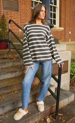 A woman stands on outdoor stone steps in front of a brick building, wearing a black and cream striped off-the-shoulder knit sweater, blue jeans, and cream slippers with gold hardware.