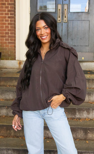 A woman with long dark hair stands on outdoor concrete steps in front of a brick building and dark metal doors, wearing a dark brown zip-front hooded jacket with voluminous balloon sleeves and light blue jeans.