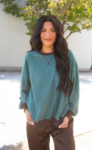 A woman wearing a teal and brown horizontally striped long-sleeve top with brown pants, standing outdoors in front of a white wall and green trees.