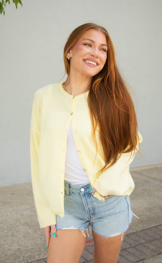 A smiling young woman with long red hair stands outdoors wearing a pale yellow cardigan over a white top and light blue denim shorts against a plain light gray wall.