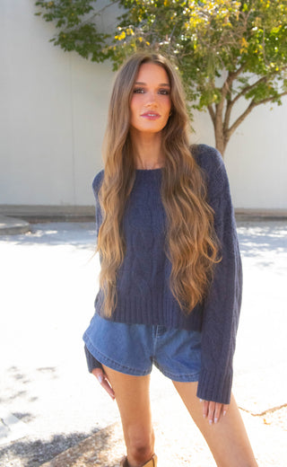 A woman wearing a dark navy blue cable-knit sweater and denim shorts, standing outdoors in front of a light-colored wall and a tree.