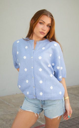 A woman wearing a light blue short-sleeve knit cardigan with white polka dots and light-wash denim shorts, standing against a plain light gray wall.