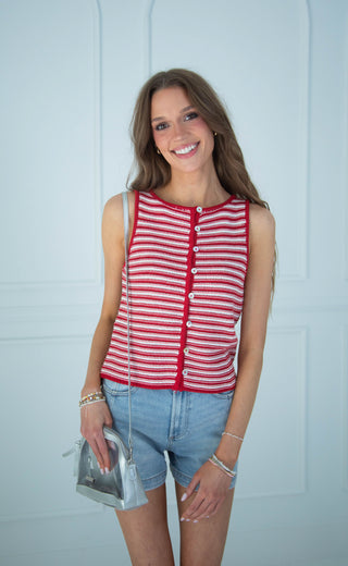 A smiling woman wearing a red and white striped sleeveless top with white buttons, paired with light blue denim shorts, holding a small silver purse against a light blue paneled wall.