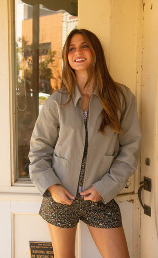 A woman smiling and posing in a light gray button-up jacket over a patterned top and dark floral shorts, standing in front of a cream-colored door.