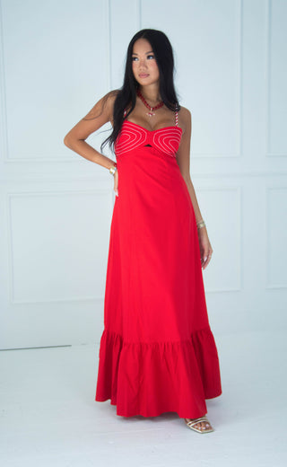 A woman modeling a bright red maxi dress with a sweetheart neckline, white curved stripe details on the bodice, and a tiered ruffle hem, set against a white paneled wall.