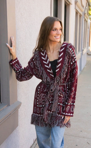 A woman wearing a burgundy and white patterned fringed cardigan leans against a beige building wall on a sidewalk.