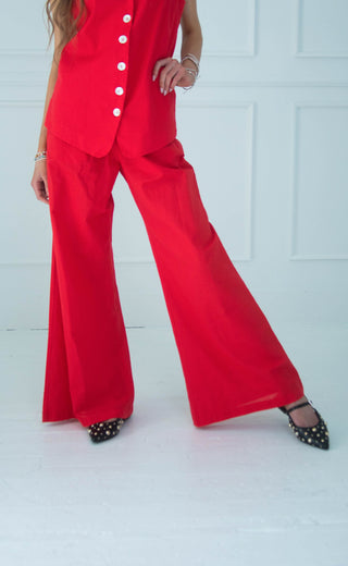 A model wearing a bright red matching two-piece set consisting of a sleeveless button-front top and wide-leg pants, paired with black pointed flats with gold studs, set against a white paneled wall.