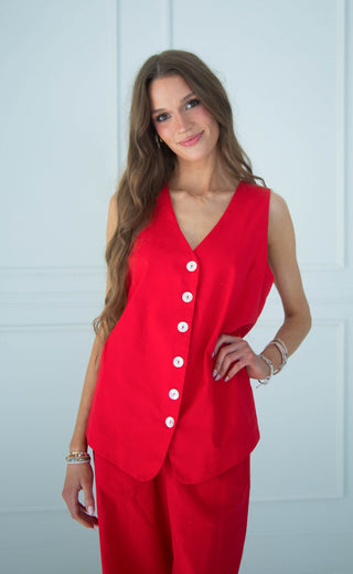 A woman modeling a bright red sleeveless vest with a V-neck and white buttons, paired with matching red pants, set against a light blue paneled wall.