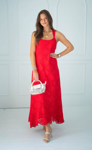 A woman modeling a bright red sleeveless eyelet maxi dress, holding a small white handbag, standing against a white paneled wall.