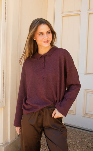 A woman wearing a long-sleeve polo-style knit top in deep plum purple and brown satin pants, standing in front of a cream-colored paneled door.