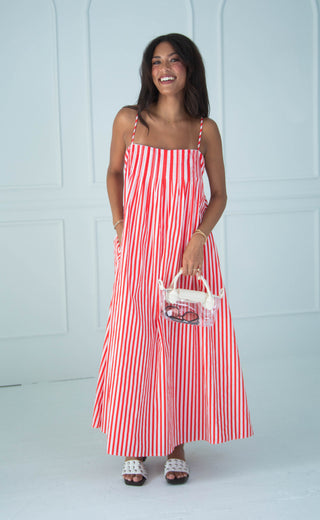 A woman modeling a red and white vertically striped maxi dress with thin straps, holding a small white handbag, set against a white paneled studio wall.