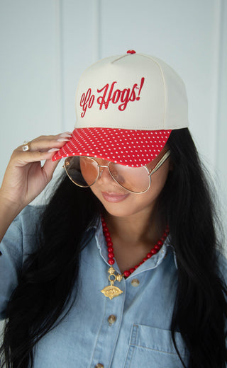 A woman wearing a cream baseball cap with red embroidered 'Go Hogs!' text and a red-and-white polka dot visor, styled with round gold-framed sunglasses, a red beaded necklace, and a light blue denim shirt.