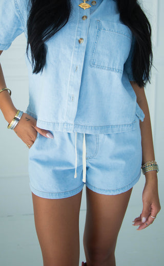 A person wearing a light blue denim matching set consisting of a short-sleeve button-front top and drawstring shorts, photographed against a plain light background.