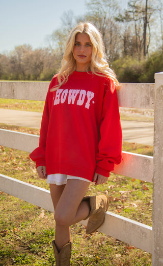 Woman wearing a red 'HOWDY' mockneck sweatshirt standing outdoors by a fence.