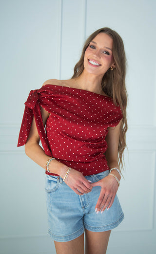 A woman modeling an off-the-shoulder red top with white polka dots and a tie detail, paired with denim shorts, against a light blue paneled wall.