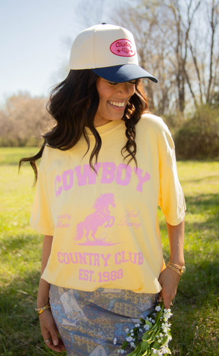 Woman wearing a yellow 'Cowboy Country Club' graphic t-shirt and white trucker hat outdoors.