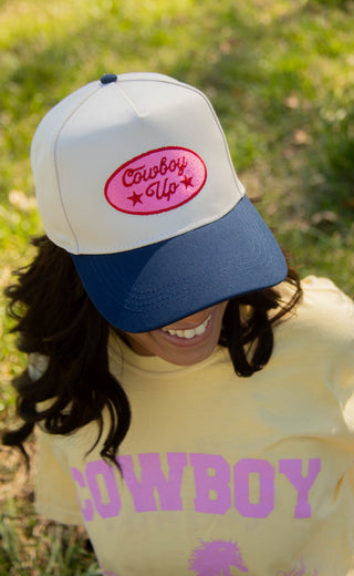 Person wearing a white and blue trucker hat with 'Cowboy Up' text, and a yellow shirt with 'Cowboy' text, outdoors.