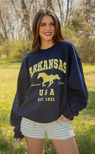 Woman wearing a navy sweatshirt with 'Arkansas USA' text and horse graphic outdoors.