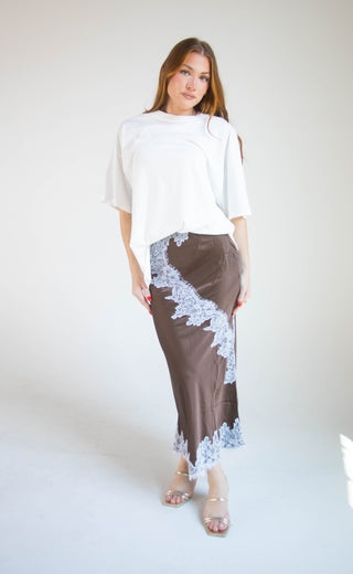 A woman modeling a white asymmetrical short-sleeve top and a brown midi skirt with light blue lace trim, standing against a plain white studio background.