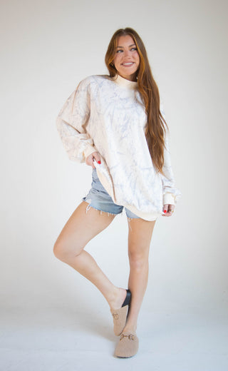 A woman posing in a cream-colored, oversized tie-dye mock-neck sweater, light-wash denim shorts, and beige slip-on shoes against a plain light gray studio background.