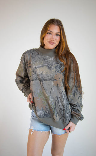 A woman modeling a long-sleeve crewneck sweatshirt with a gray and brown camouflage pattern, paired with light blue denim shorts, against a plain light gray background.