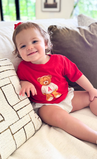 A baby girl sitting on a beige couch wearing a red short-sleeve t-shirt with a teddy bear graphic and white diaper. The background shows a cozy living room setting with pillows.