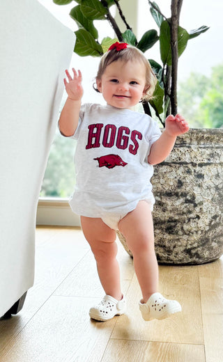A baby toddler wearing a white short-sleeve bodysuit with 'HOGS' and a hog graphic in red, standing on a light wood floor next to a large potted plant.