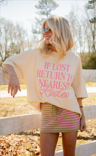 Woman wearing a light-colored oversized graphic t-shirt with "if lost return to nearest rodeo" and a striped skirt outdoors. 