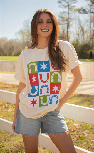 Woman wearing a colorful graphic t-shirt with horseshoes outdoors near a white fence.