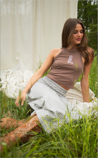Woman sitting in grass wearing a brown top and checkered skirt.
