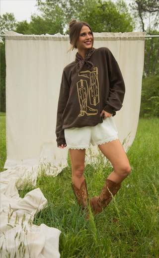 Woman in a brown sweatshirt with a graphic design, white shorts, and brown boots standing in a grassy area.