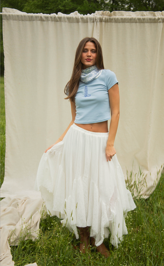 Woman wearing a light blue top and white skirt standing in a grassy area with a white curtain in the background.