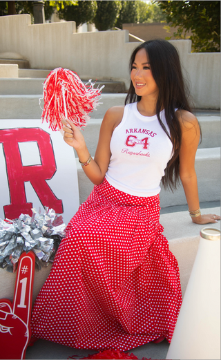 A woman posing on outdoor concrete steps wearing a white tank top with 'ARKANSAS 64 Razorbacks' printed in red, paired with a long red and white polka dot skirt. 