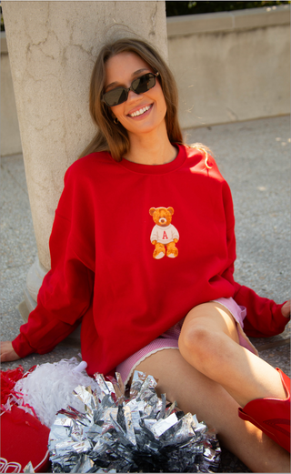 A woman wearing a bright red crewneck sweatshirt with a teddy bear graphic sits on a stone ledge outdoors. The sweatshirt features a small orange-brown teddy bear wearing a white shirt with the letter 'A' on the chest.