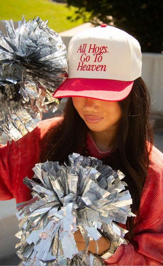 A woman outdoors wearing a white and red baseball cap with the text 'All Hogs Go To Heaven' in red, holding silver foil pom-poms. She is wearing a red sweatshirt.