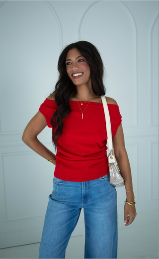 Women wearing a red boat neck asymmetrical top with light wash jeans and a clear purse.