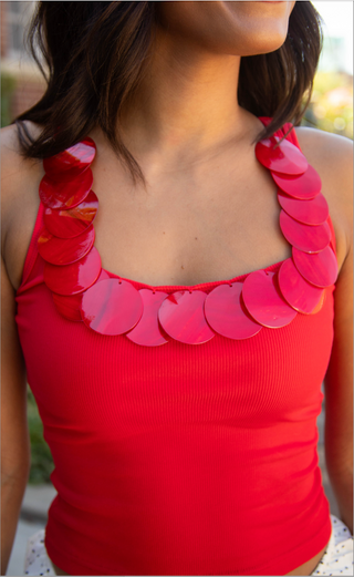 A close-up of a woman wearing a bright red ribbed tank top with glossy, overlapping circular shell-like embellishments around the scoop neckline.