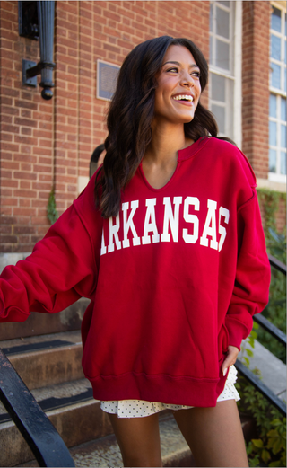 A woman smiling on outdoor brick steps wearing a red crewneck sweatshirt with 'ARKANSAS' printed in white arched letters across the chest.