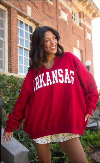A woman smiling outdoors in front of a brick building, wearing an oversized red sweatshirt with 'ARKANSAS' printed in white across the chest.