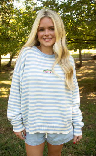 A smiling blonde woman standing outdoors in a grassy wooded area, wearing a light blue and white horizontally striped long-sleeve crewneck sweatshirt with a small embroidered alligator graphic on the left chest, paired with denim shorts.