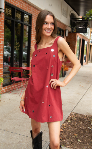 A woman stands on a city sidewalk wearing a rust-red sleeveless mini dress with asymmetrical silver and black embellishments. The background shows a brick storefront with windows and outdoor seating.