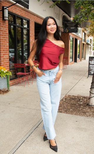 A woman posing on a sidewalk in front of a brick storefront, wearing a metallic red one-shoulder top, light wash blue jeans, and black heels.
