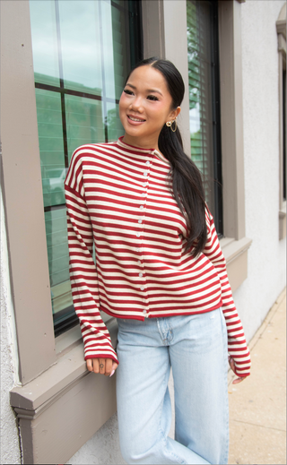 A woman wearing a red and white striped long-sleeve knit top with white buttons, paired with light blue jeans, standing outdoors next to a window.