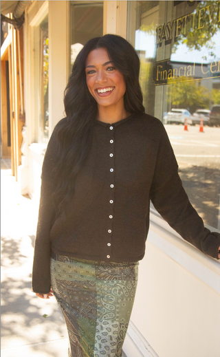 A smiling woman with long dark hair stands outdoors in front of a storefront window, wearing a dark brown button-front cardigan and a green paisley patterned skirt.
