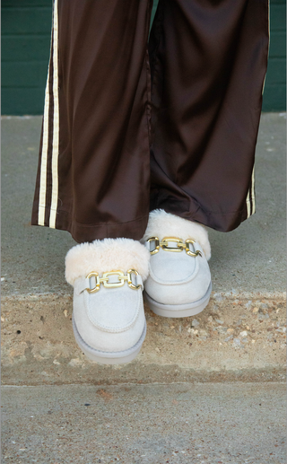 A close-up of a person's feet wearing light gray slip-on loafers with fluffy white shearling lining and gold chain details, paired with brown satin track pants with white stripes.